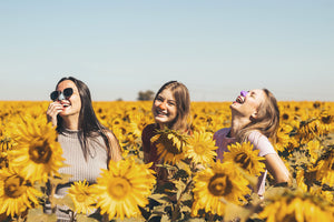 Three young girls laughing in a field of sunflowers, wearing colored sunscreen for nose.